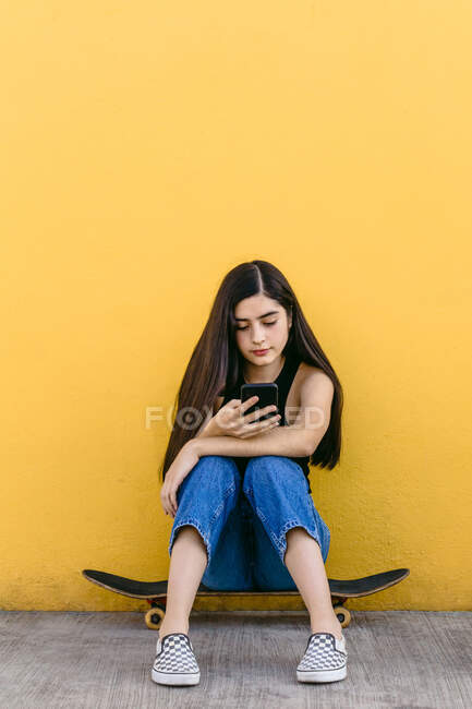 Young female skater surfing internet on cellphone while sitting on skateboard on walkway in daytime — Stock Photo