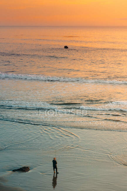 Desde arriba increíble paisaje con solitario pescador irreconocible en la orilla de arena húmeda lavado por las olas de espuma del océano bajo el cielo colorido durante la puesta del sol - foto de stock