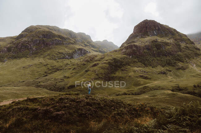 Vue arrière de l'homme méconnaissable debout sur une colline herbeuse rugueuse pendant le voyage à travers Glencoe dans la campagne britannique par temps nuageux — Photo de stock