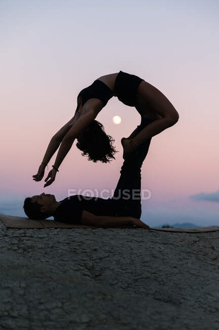 Vista lateral de silueta de mujer flexible haciendo backbend y balanceo sobre las piernas del hombre durante sesión de acroyoga contra puesta de sol cielo con luna - foto de stock