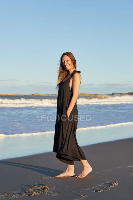 Vue latérale de la femme souriante en robe d'été debout sur le bord de mer sablonneux et regardant la caméra — Photo de stock