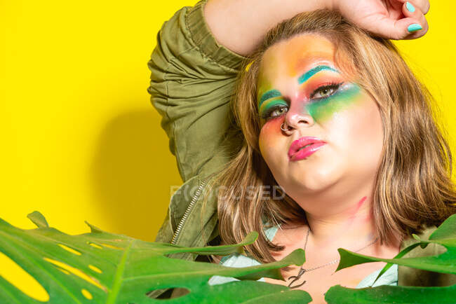 Пухка молода жіноча модель з барвистим креативним макіяжем торкаючись голови і дивлячись на камеру на жовтому тлі — стокове фото