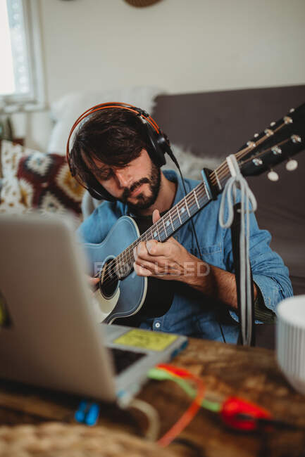 Молодий чоловік в навушниках грає на гітарі біля столу з ноутбуком і синтезатором вдома дивлячись на камеру — стокове фото