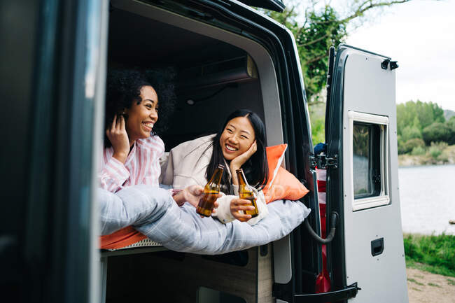 Alegre joven multirracial mujeres beber cerveza mientras se enfrían juntos en autocaravana durante el viaje de verano - foto de stock
