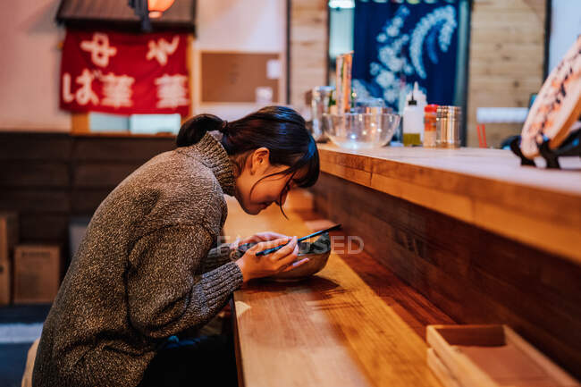 Vue latérale de femme asiatique heureuse en tenue décontractée tout en étant assis au comptoir avec baguettes et bol avec ramen dans le café — Photo de stock