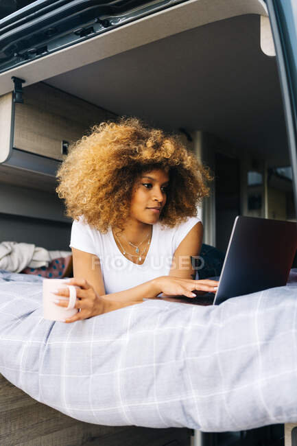 Оптимистичная черная женщина с кружкой горячего напитка улыбается и просматривает социальные сети в нетбуке, лежа на кровати в фургоне и отдыхая утром — стоковое фото