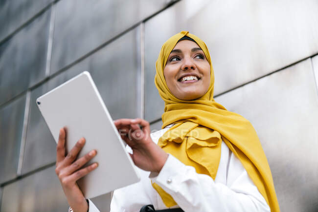 Empreendedora muçulmana sorridente em hijab amarelo em pé na rua e tablet de navegação — Fotografia de Stock
