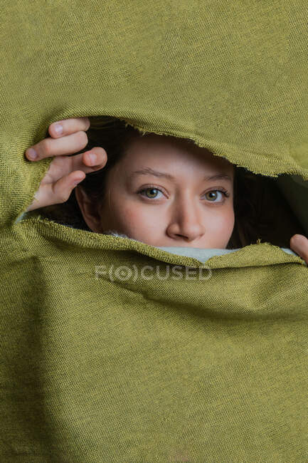 Giovane donna dagli occhi verdi guardando la fotocamera mentre si nasconde dietro un panno verde strappato — Foto stock