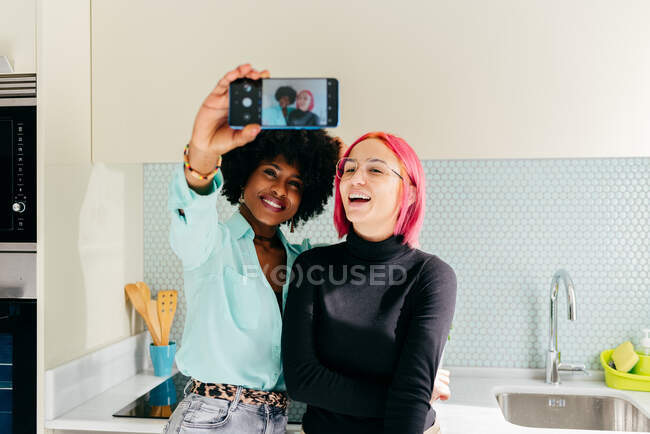 Young stylish diverse female friends in casual clothes smiling while taking selfie with mobile phone at home kitchen — Stock Photo