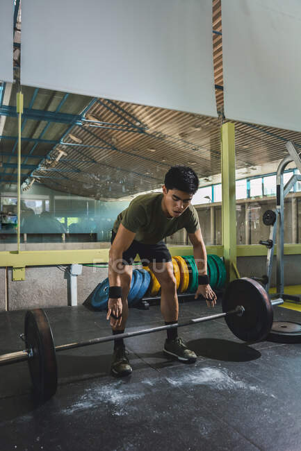 Сосредоточенный азиатский спортсмен делает тягу с тяжелым штангой во время тренировки в тренажерном зале глядя вниз — стоковое фото