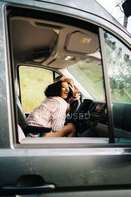 Vista lateral do romântico jovem viajante afro-americano alegre sentado no assento do motorista e olhando para a câmera enquanto desfruta da viagem de verão na natureza — Fotografia de Stock