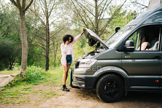 Young African American female traveler standing near camper van with opened hood and talking with repair service by mobile phone while getting troubles during summer trip in nature — Stock Photo