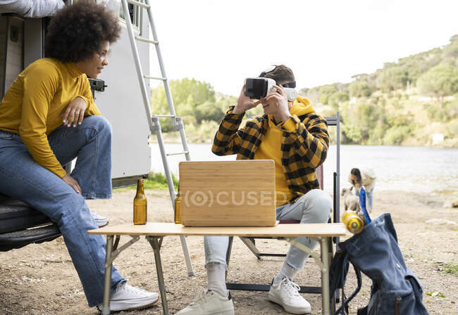 Веселый человек в очках виртуальной реальности исследует виртуальную реальность рядом с восхитительной черной девушкой, отдыхая на природе вместе — стоковое фото