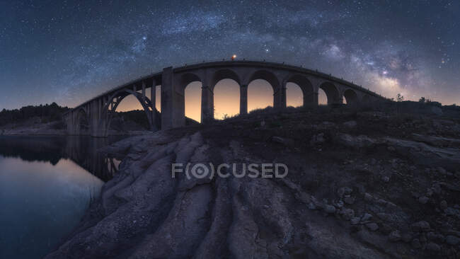 Increíble paisaje de envejecido puente de piedra con elementos arqueados que cruzan el río bajo el cielo nocturno con la brillante Vía Láctea y la luz del atardecer - foto de stock