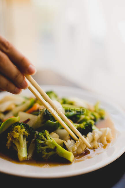 Hand with chopsticks lifting piece from white ceramic plate with broccoli and squid meal — Stock Photo