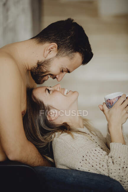 Glückliche Männer und Frauen mit einem Becher Heißgetränk, die einander lächelnd anschauen, während sie morgens Zeit zu Hause verbringen — Stockfoto