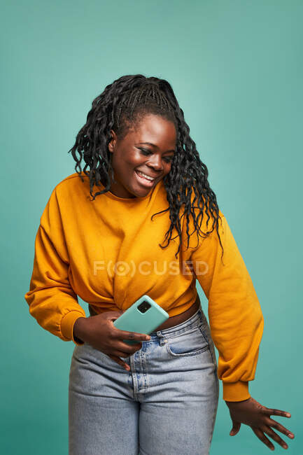 Femme afro-américaine souriante en jeans et pull jaune tenant un smartphone moderne et dansant contre un mur bleu en studio — Photo de stock