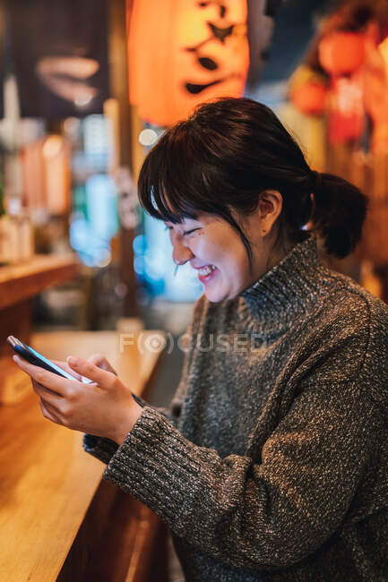 Asian lady in casual sweater smiling while using mobile phone at counter in traditional ramen bar — Stock Photo