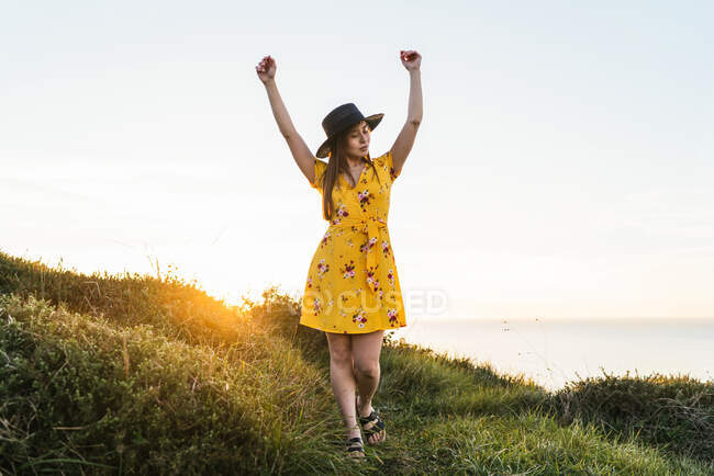 Longitud completa atractiva joven hembra en vestido amarillo y sombrero de pie con los brazos levantados en el verde prado herboso en el campo soleado - foto de stock