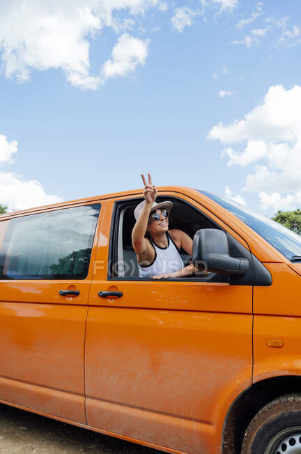 Cheerful female traveler peeping out of van window and showing two fingers gesture while enjoying road trip in summer — Stock Photo