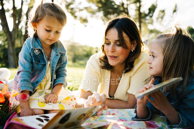 Счастливая молодая мама с маленькими дочками, лежащими на одеяле и играющими в игры, проводя летний день вместе в солнечном парке — стоковое фото