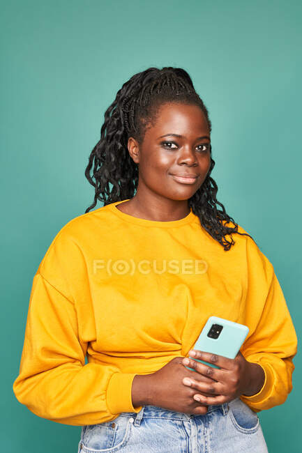 Afroamerikanerin in Jeans und gelbem Pullover hält modernes Smartphone vor blauer Wand im Studio — Stockfoto