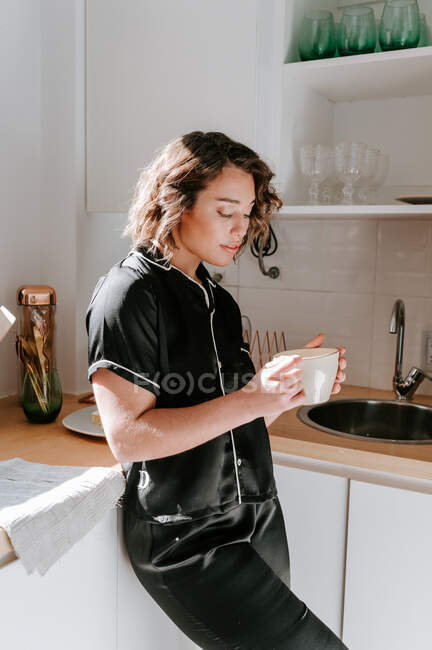 Вид сбоку нежной женщины в пижаме, стоящей на кухне с чашкой напитка во время завтрака по утрам — стоковое фото