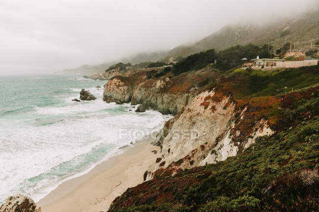 High angle of wonderful scenery of stormy ocean water with foam waves washing sand beach against foggy highland shoreline under gray sky in Big Sur — Stock Photo