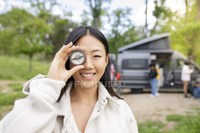 Heureuse femme asiatique souriante et regardant la caméra tout en tenant boussole vintage près des yeux pendant le voyage à la campagne avec des amis — Photo de stock