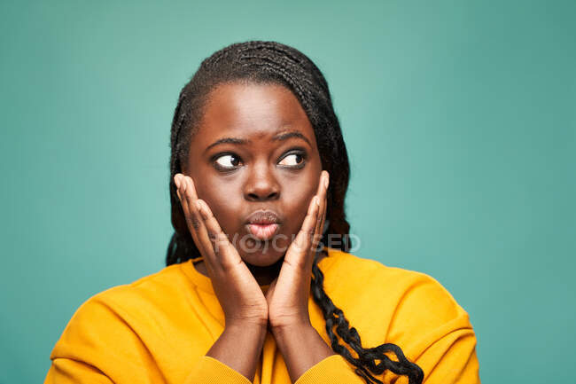 Glad African American female in yellow clothes pouting lips and looking away while holding face in hands against blue background — Stock Photo