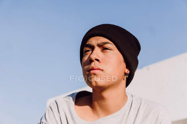 From below confident ethnic teenager and beanie in cool clothes standing in city street and looking away — Stock Photo