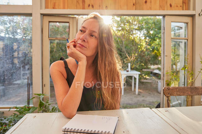 Baixo ângulo de jardineiro feminino alegre sentado à mesa e escrevendo em bloco de notas em estufa — Fotografia de Stock