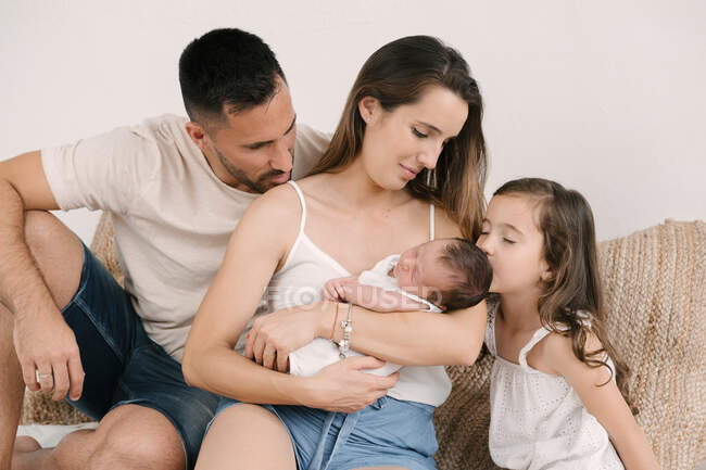 Linda niña besando al bebé recién nacido mientras está sentada en el sofá con los padres en casa - foto de stock