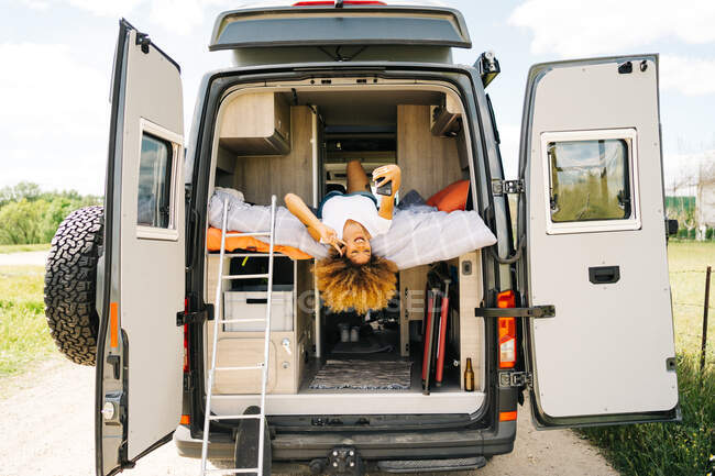 Positive black woman with Afro hairstyle lying upside down on bed in trailer and browsing mobile phone on sunny summer day — Stock Photo