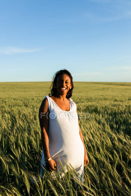 Smiling young black lady in white summer dress strolling on green wheat field while looking at camera in daytime under blue sky — Stock Photo