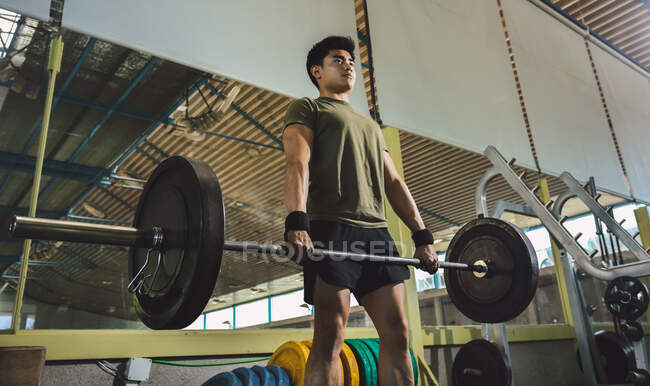 Atleta masculino asiático enfocado haciendo deadlift con barra pesada durante el entrenamiento en el gimnasio - foto de stock