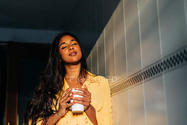 Low angle of young Hispanic female in yellow shirt closing eyes and enjoying hot beverage while leaning on wall and resting in morning at home — Stock Photo