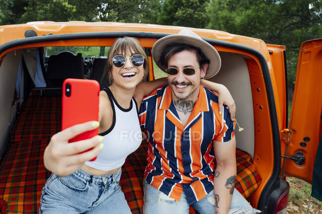 Entzücktes Paar von Reisenden, die im Lieferwagen sitzen und sich mit dem Smartphone fotografieren, während sie sich umarmen und das Sommerabenteuer genießen — Stockfoto