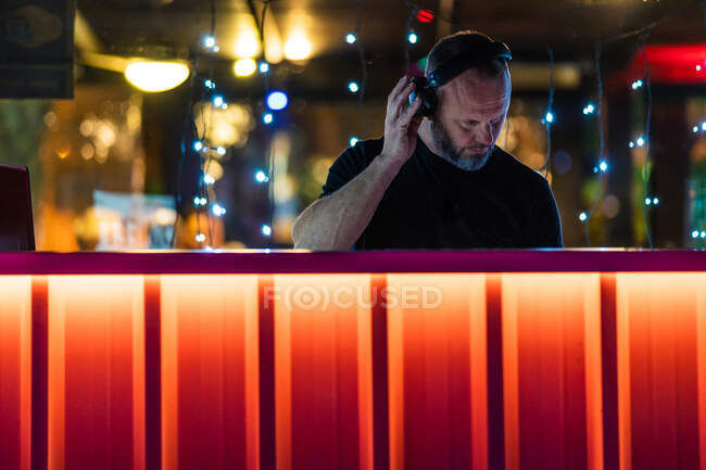 Low angle of serious middle aged man in wireless headphones concentrating and playing DJ controller while performing at concert at vibrant orange counter on stage in modern nightclub — Stock Photo