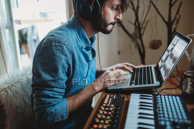 Side view of young man in headphones using synthesizer and laptop at table at home — Stock Photo