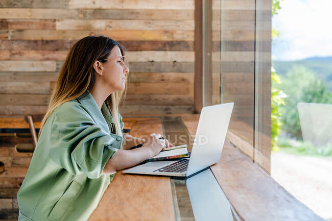 Side view of concentrated adult lady in trendy outfit and eyeglasses sitting in chair at table while writing notes in notepad next to open netbook in light cafe near window — Stock Photo