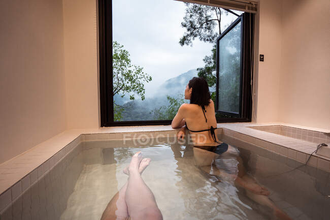 Безликі люди в купальнику в ванні онсена на курорті поруч з вікном з видом на пагорби і зелені дерева — стокове фото
