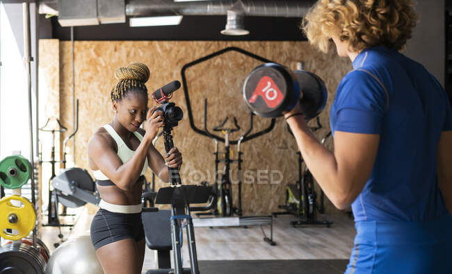 Ethnic female athlete recording video of crop unrecognizable sportsman with dumbbell on photo camera in gym — Stock Photo
