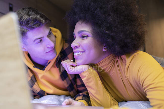 Щасливий багаторасовий чоловік і жінка посміхаються відпочиваючи і переглядаючи ноутбук в наметі вночі — стокове фото