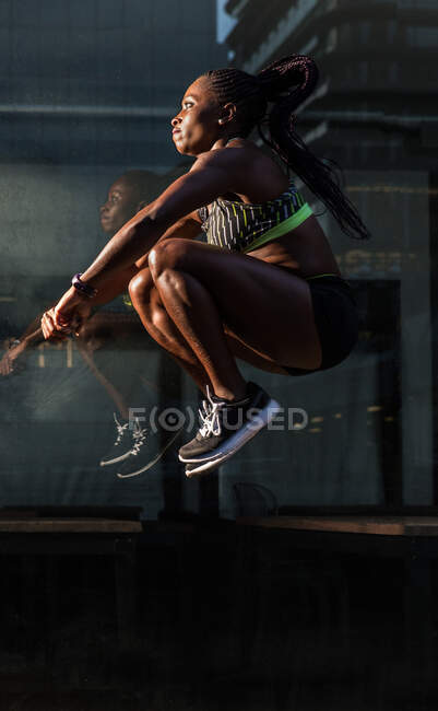 Vista lateral de una mujer negra fuerte saltando alto cerca de la pared de vidrio del edificio moderno mientras hace ejercicio en la calle de la ciudad en un día soleado - foto de stock