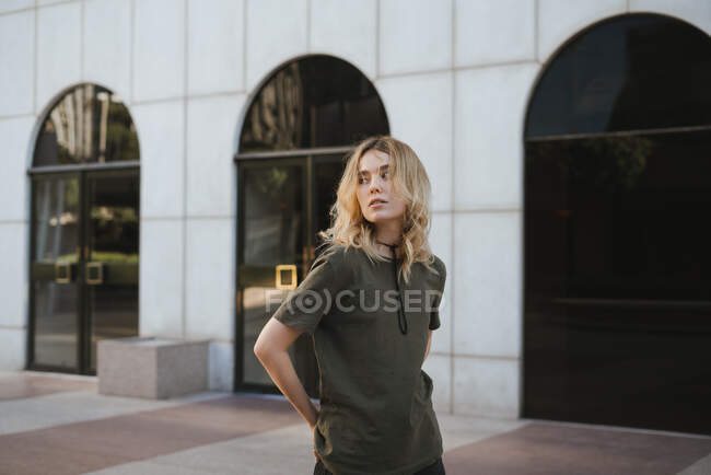 Молода блондинка в повсякденному одязі стоїть з схрещеними ногами на прогулянці, дивлячись на камеру в місті — стокове фото