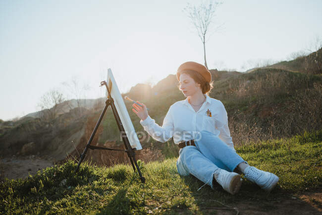 Corpo inteiro de jovem mulher em boina e roupas elegantes sentadas em gramado perto de colinas enquanto pintava imagem com escova em tela no cavalete em dia ensolarado sob o céu — Fotografia de Stock