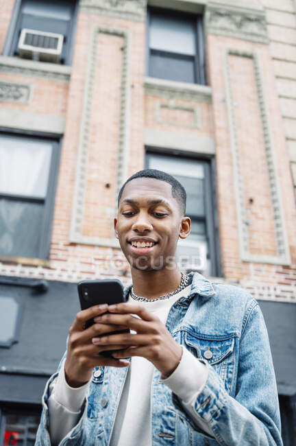 Von unten positiver schwarzer junger Mann im Jeans-Outfit, der beim Spazierengehen in der Stadt per Handy Nachrichten übermittelt — Stockfoto