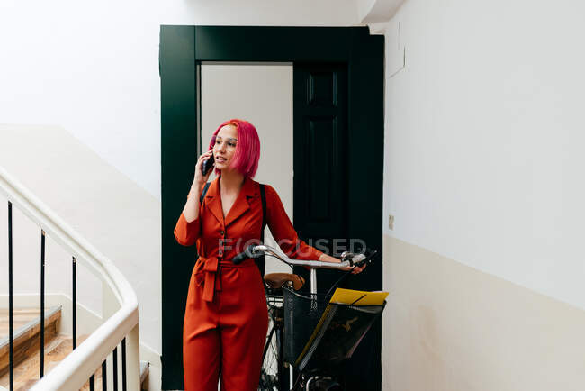 Joven mujer con estilo en traje rojo con mochila hablando en el teléfono inteligente mientras está de pie con la bicicleta en la escalera - foto de stock