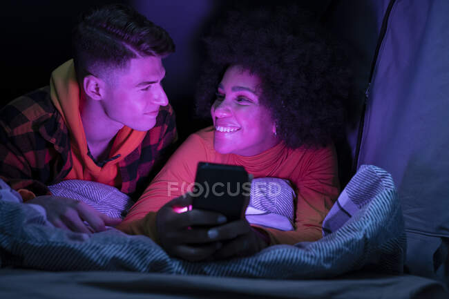 Щасливий багаторасовий чоловік і жінка посміхаються і дивляться один на одного, відпочиваючи і переглядаючи мобільний телефон в наметі вночі — стокове фото